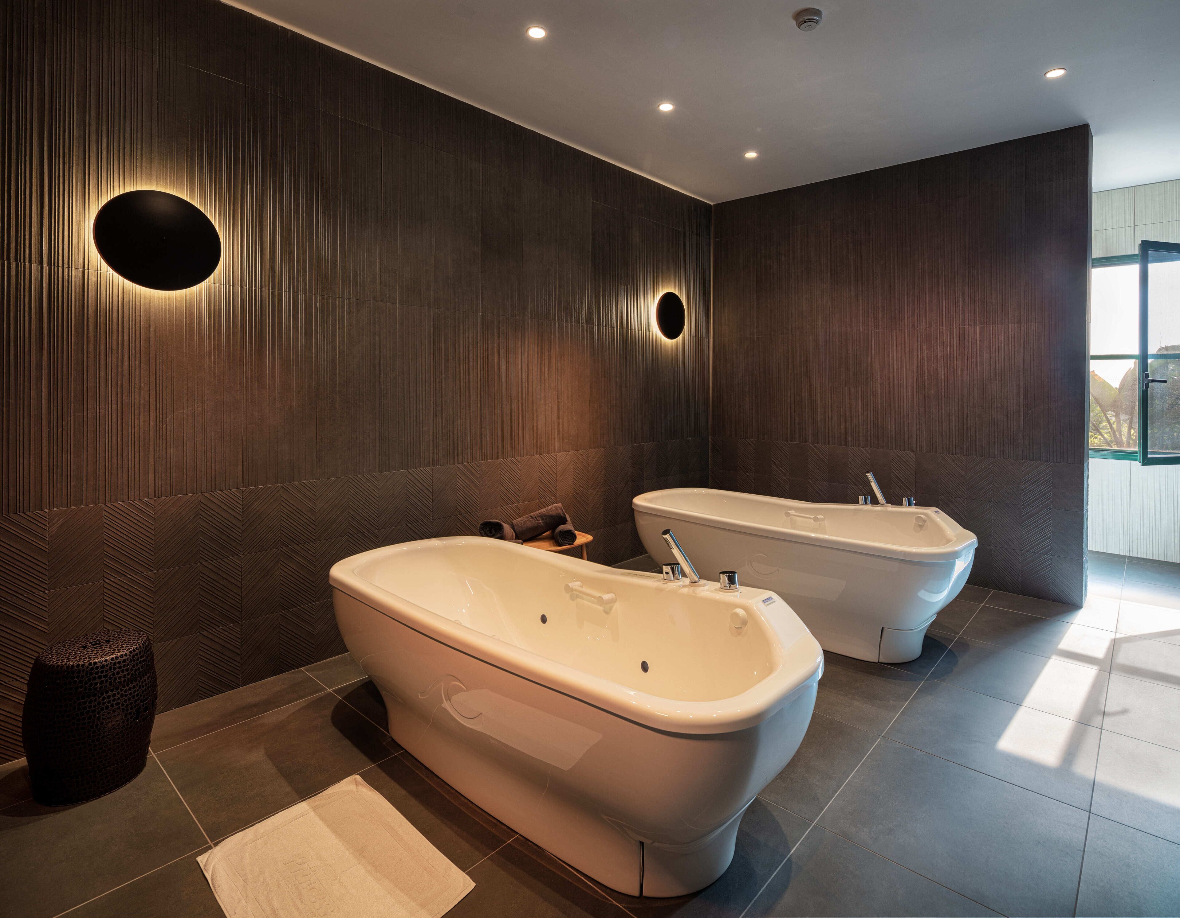 A modern bathroom with two freestanding tubs, a round mirror, and a textured wall.