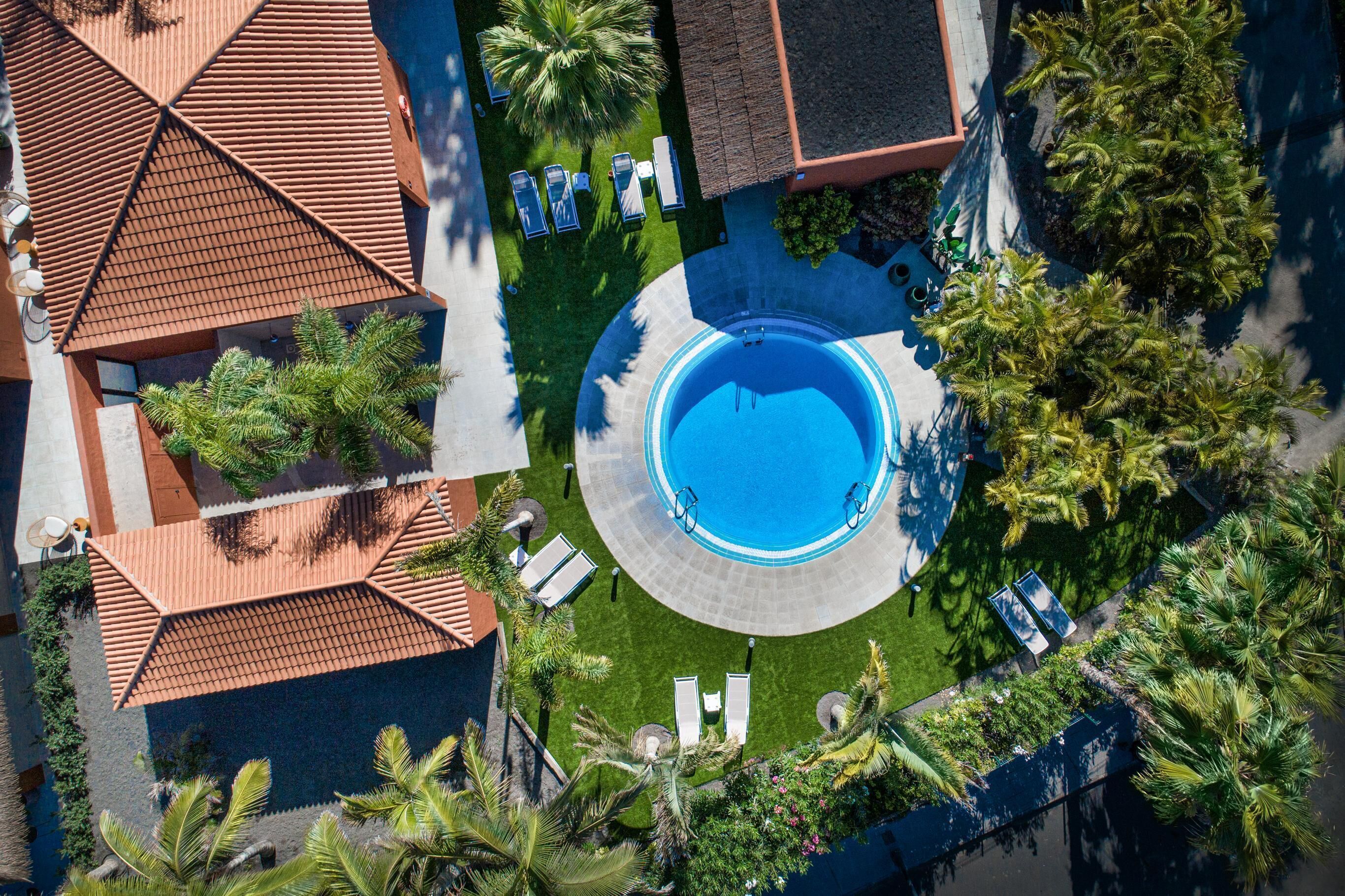 Vista aérea de un complejo turístico con piscina central, sillas de descanso y palmeras.