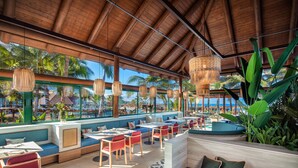 A spacious outdoor dining area with wooden tables and red chairs, overlooking a pool and palm trees.