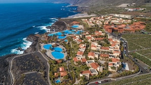 An aerial view of a coastal resort with multiple swimming pools, beachfront villas, and a mountainous backdrop.