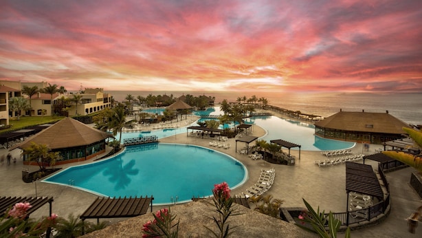 A resort with a large swimming pool, multiple lounge areas, and a view of the ocean during sunset.