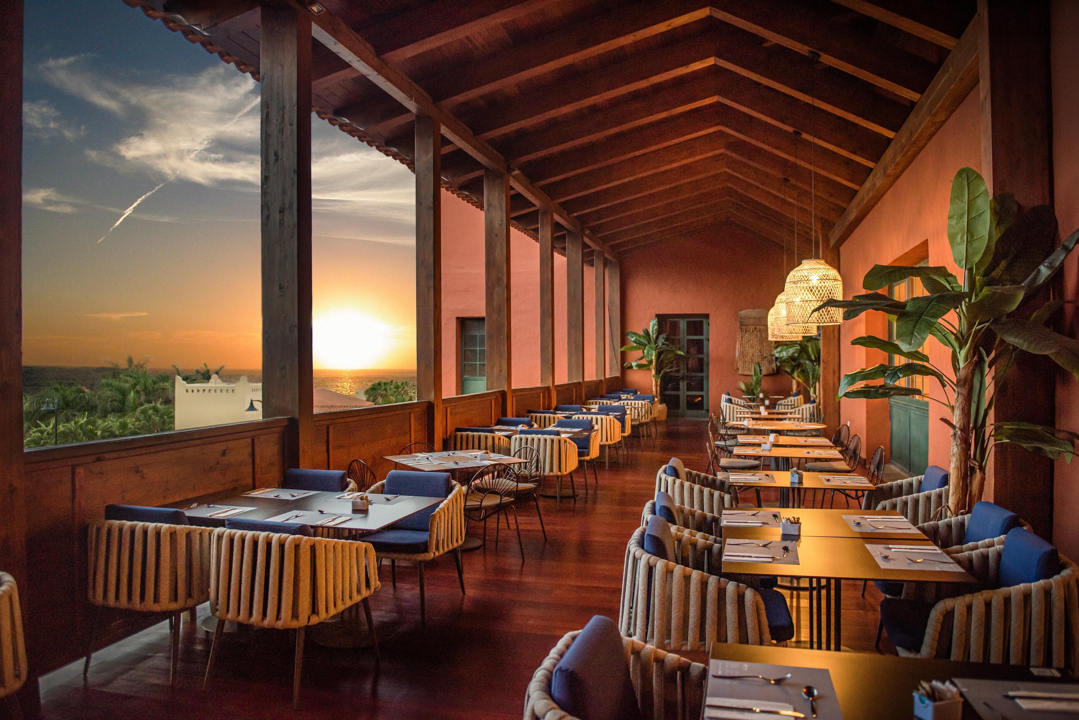 A restaurant with a long table layout, large windows, and a view of the sunset.