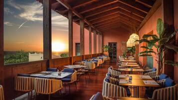 A restaurant with a long table layout, large windows, and a view of the sunset.
