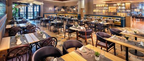 A spacious restaurant with multiple dining tables and chairs, large hanging lights, and a view of the outdoors through large windows.