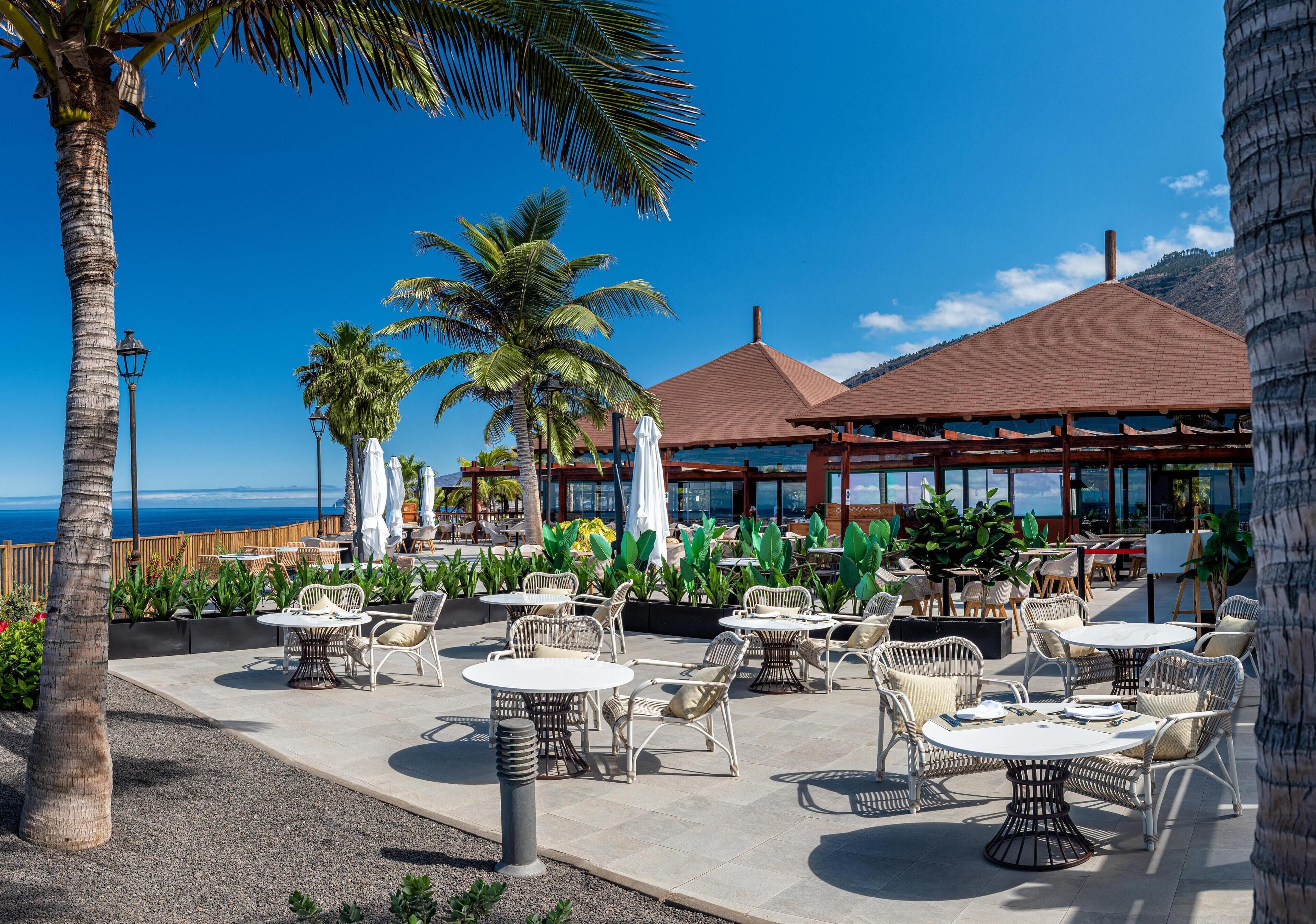 Un patio al aire libre con mesas redondas y sillas de mimbre, con vista a una playa con palmeras y un cielo azul despejado.