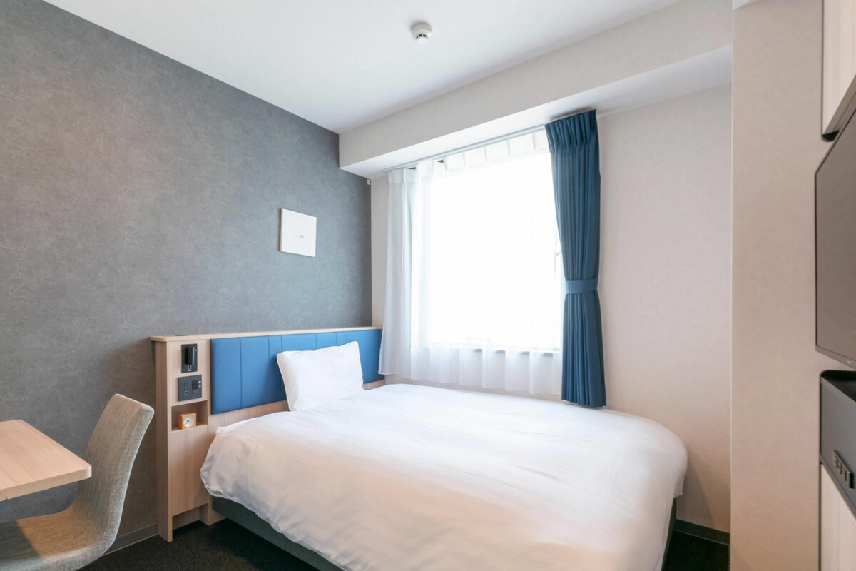 A hotel room with a bed, a desk, a chair, a TV, and a window with curtains.