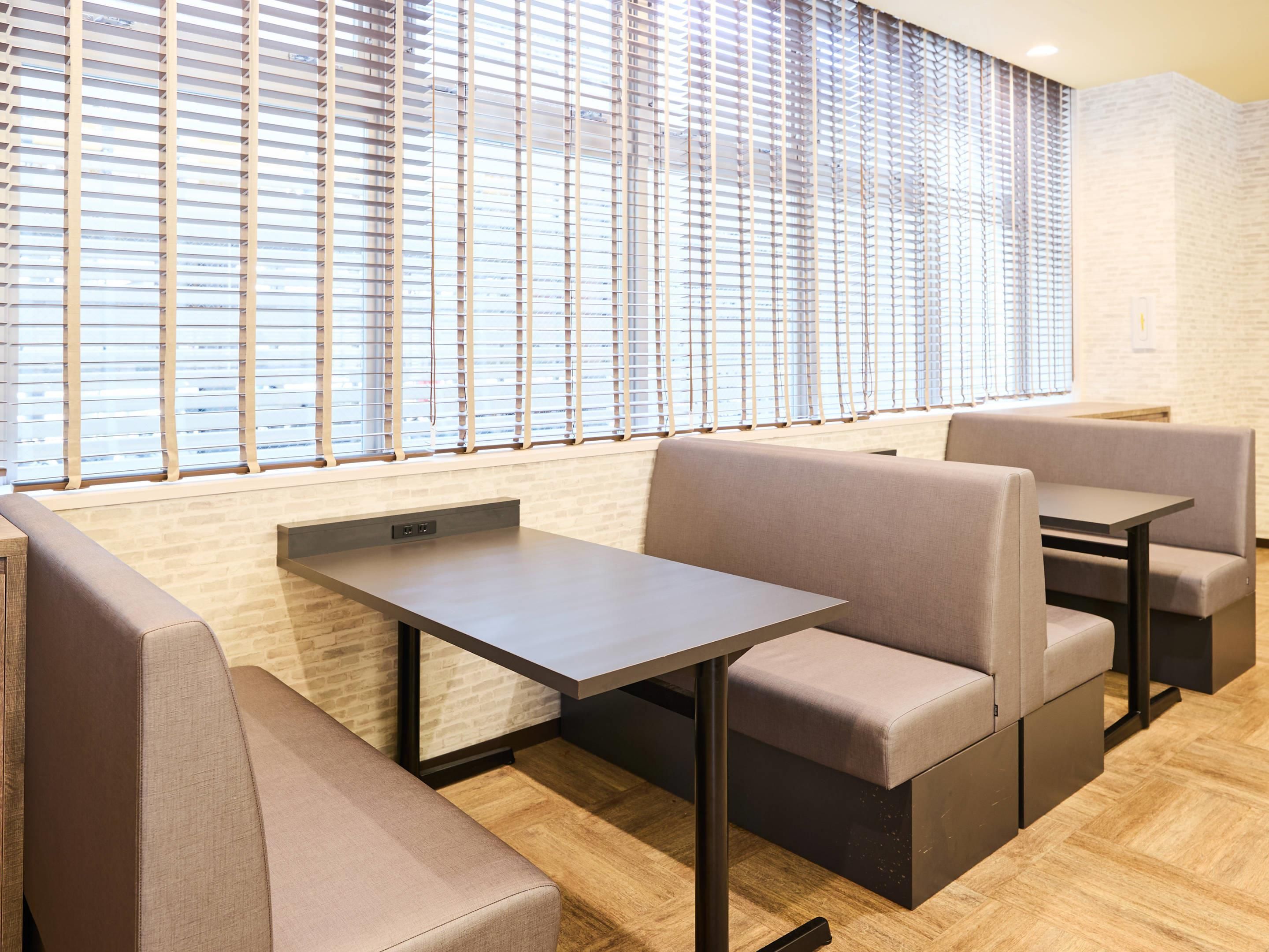 A modern dining area with cushioned booths and a long table, large windows with blinds, and a brick wall.