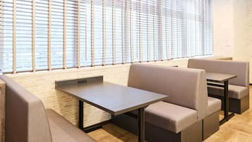 A modern dining area with cushioned booths and a long table, large windows with blinds, and a brick wall.