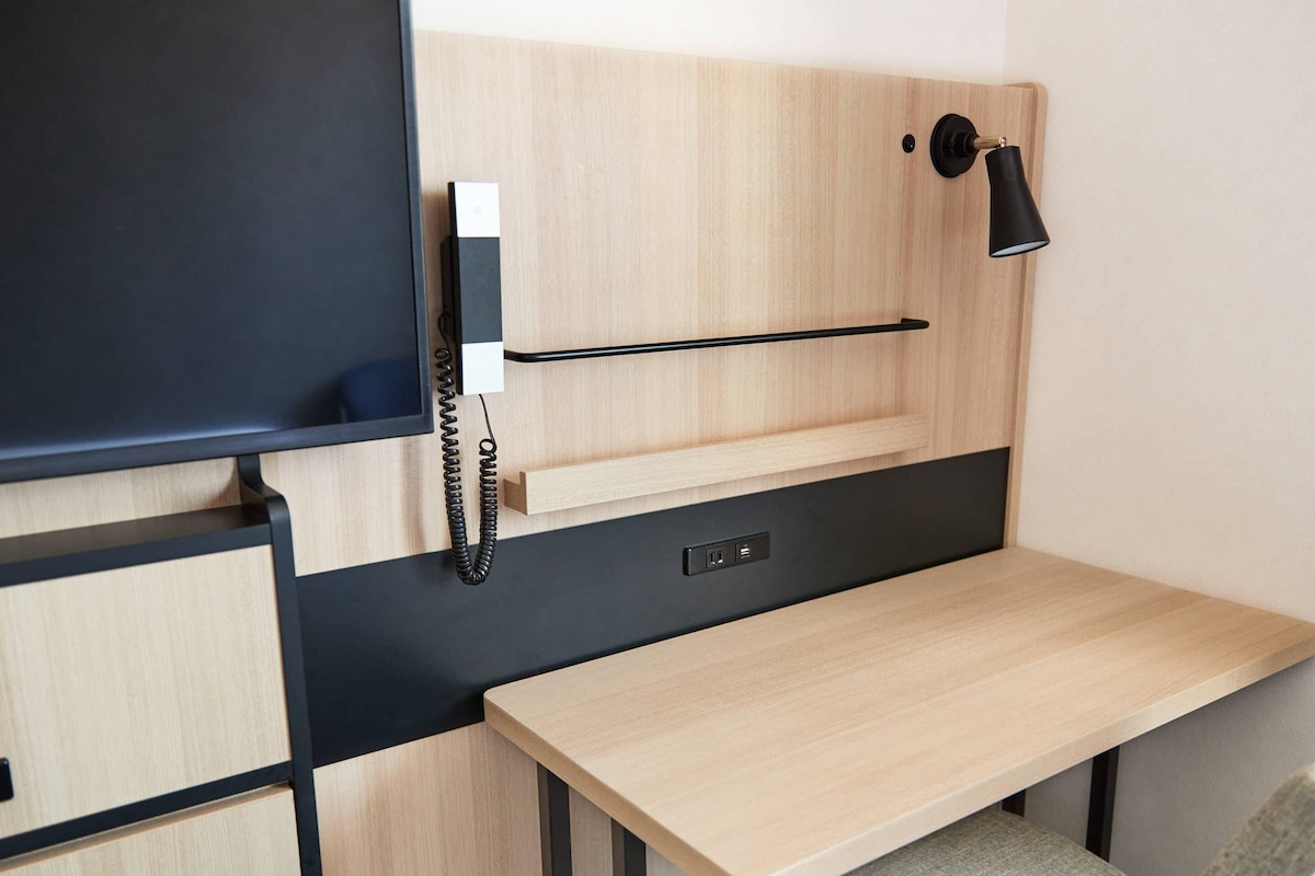 A compact room with a desk, a mounted telephone, and a wall-mounted lamp.