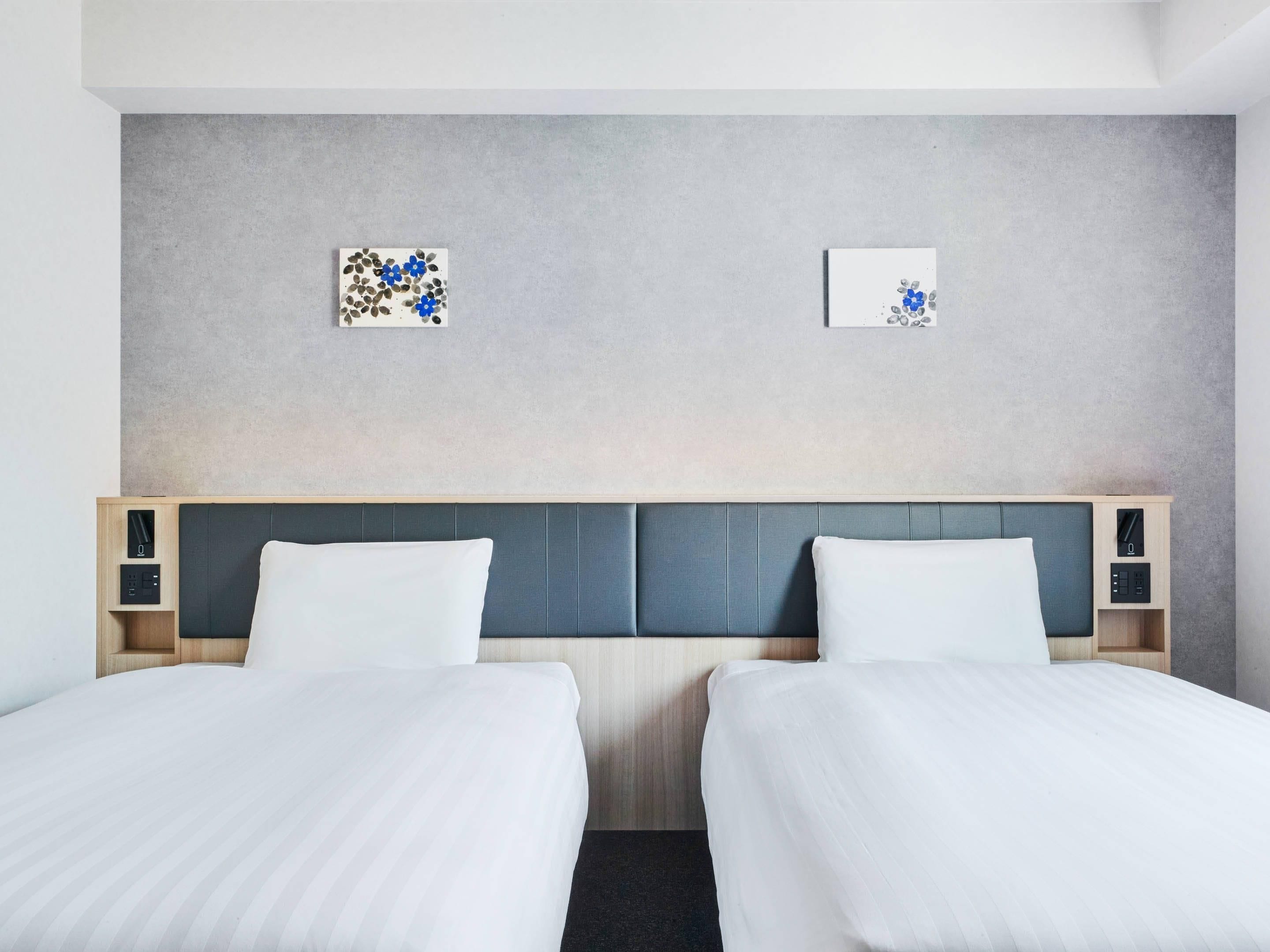 A hotel room with two beds, a grey headboard, and two bedside tables with lamps.
