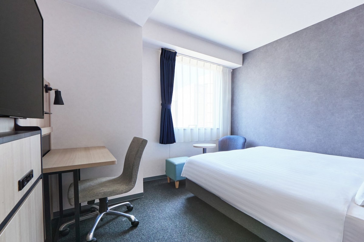 A hotel room with a bed, a desk, a chair, a TV, and a window with curtains.