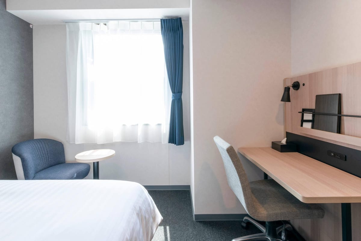 Une chambre d’hôtel avec un lit, un bureau, une chaise, une petite table et une fenêtre avec des rideaux.