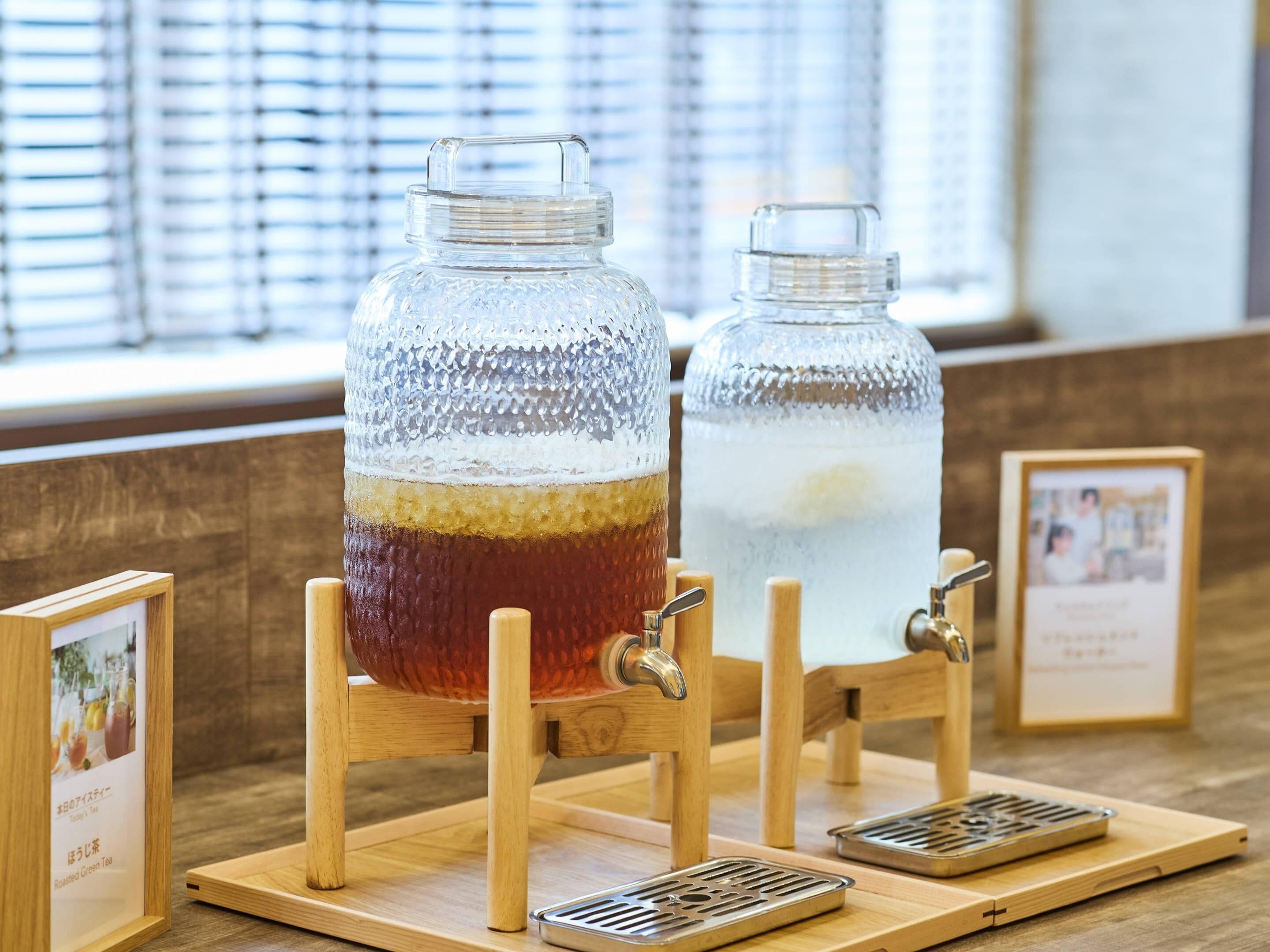 Two glass jars on wooden stands with taps, likely for serving beverages, on a wooden table with a window in the background.