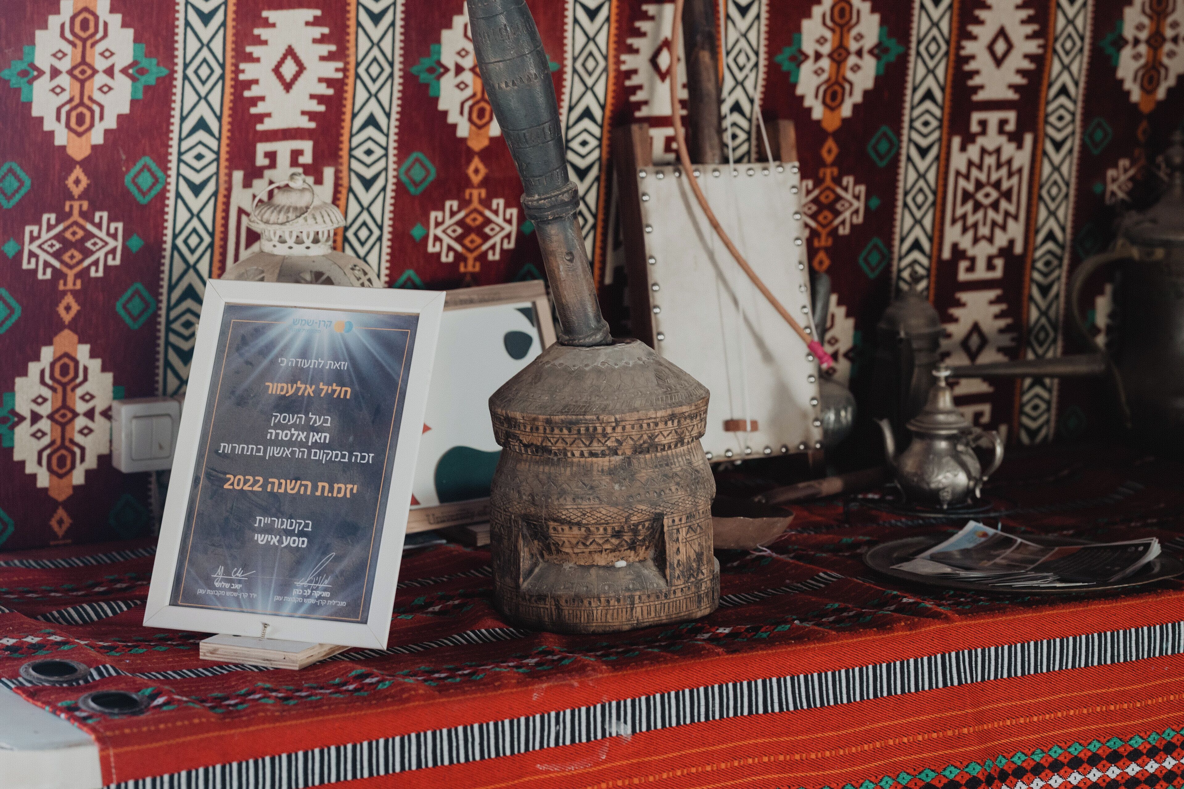 A traditional clay water pot with a metal spout, a framed certificate, and a patterned textile backdrop.