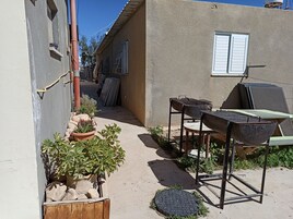 Un patio exterior estrecho con una parrilla, una mesa de madera y plantas en macetas.
