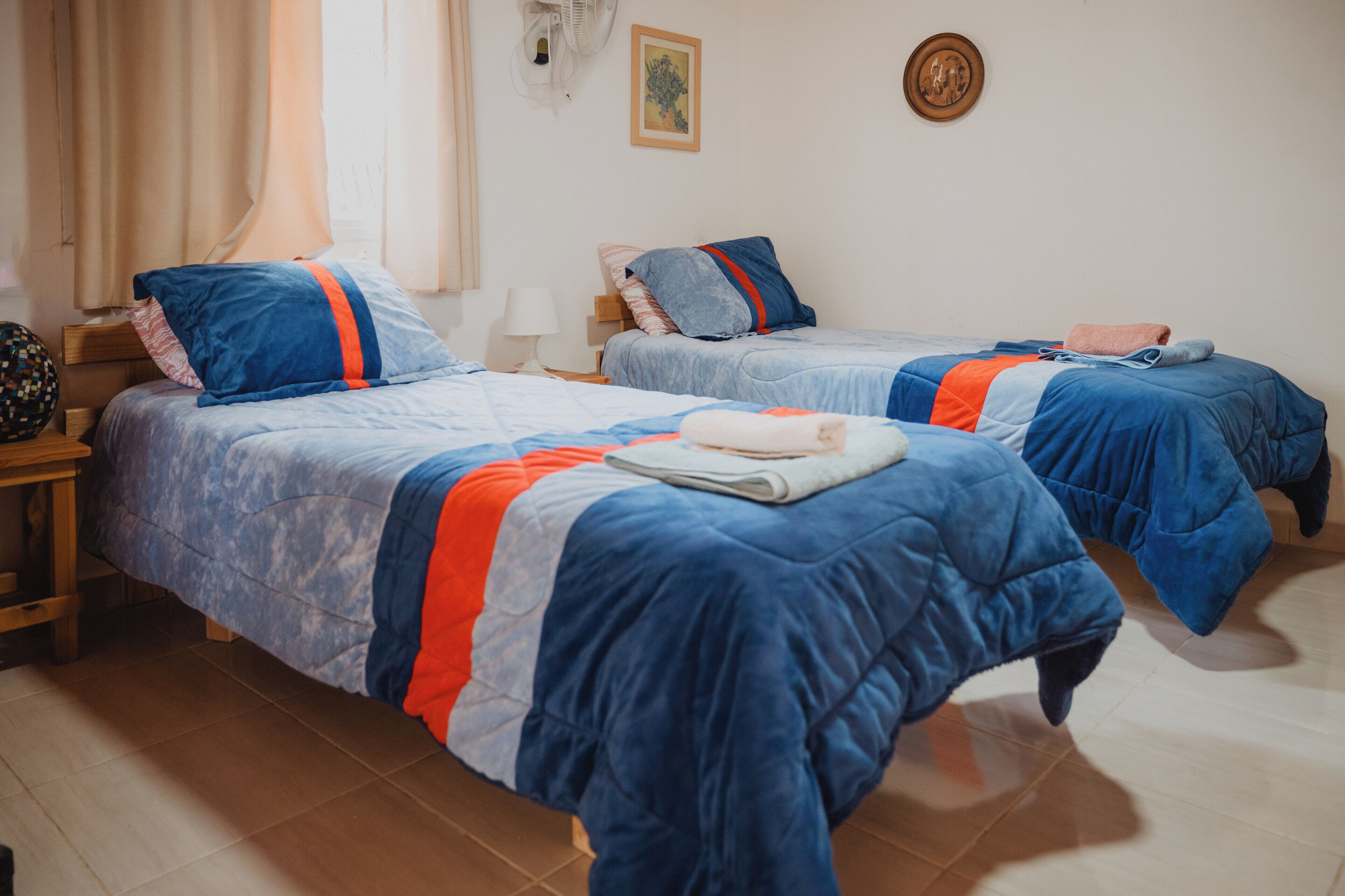 Habitación de hotel con dos camas, cada una con ropa de cama azul y detalles naranja, una mesita de noche de madera, una lámpara y un ventilador montado en la pared.