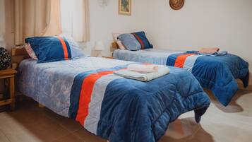Habitación de hotel con dos camas, cada una con ropa de cama azul y detalles naranja, una mesita de noche de madera, una lámpara y un ventilador montado en la pared.