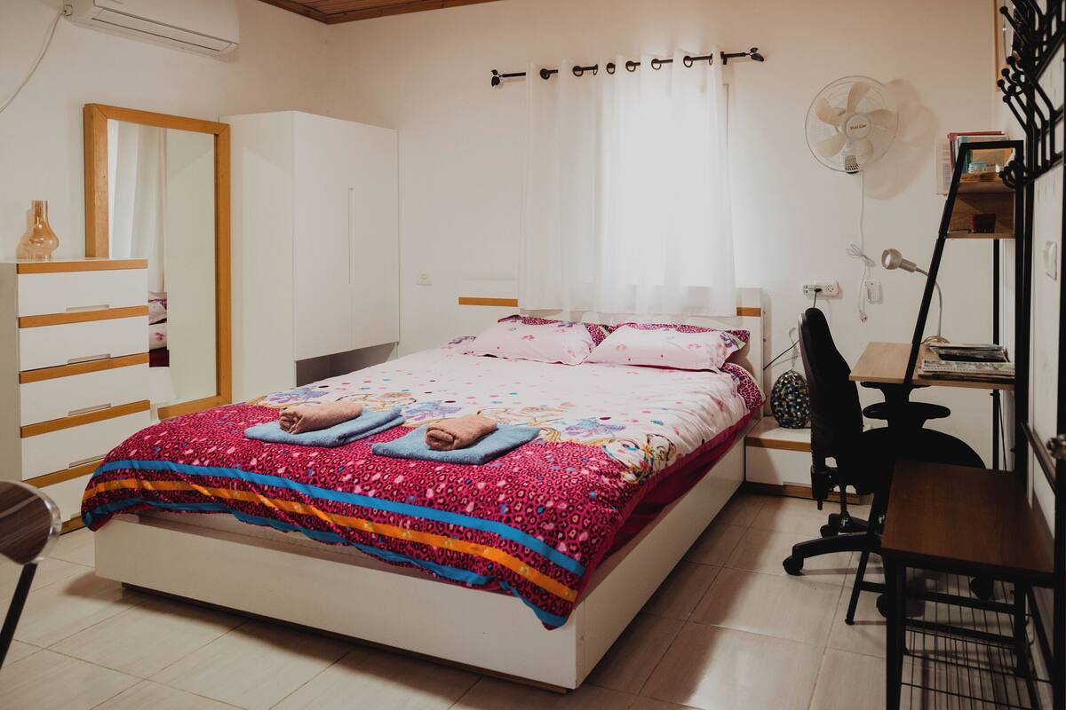 A bedroom with a bed, a desk, a chair, a mirror, a fan, and a wardrobe.