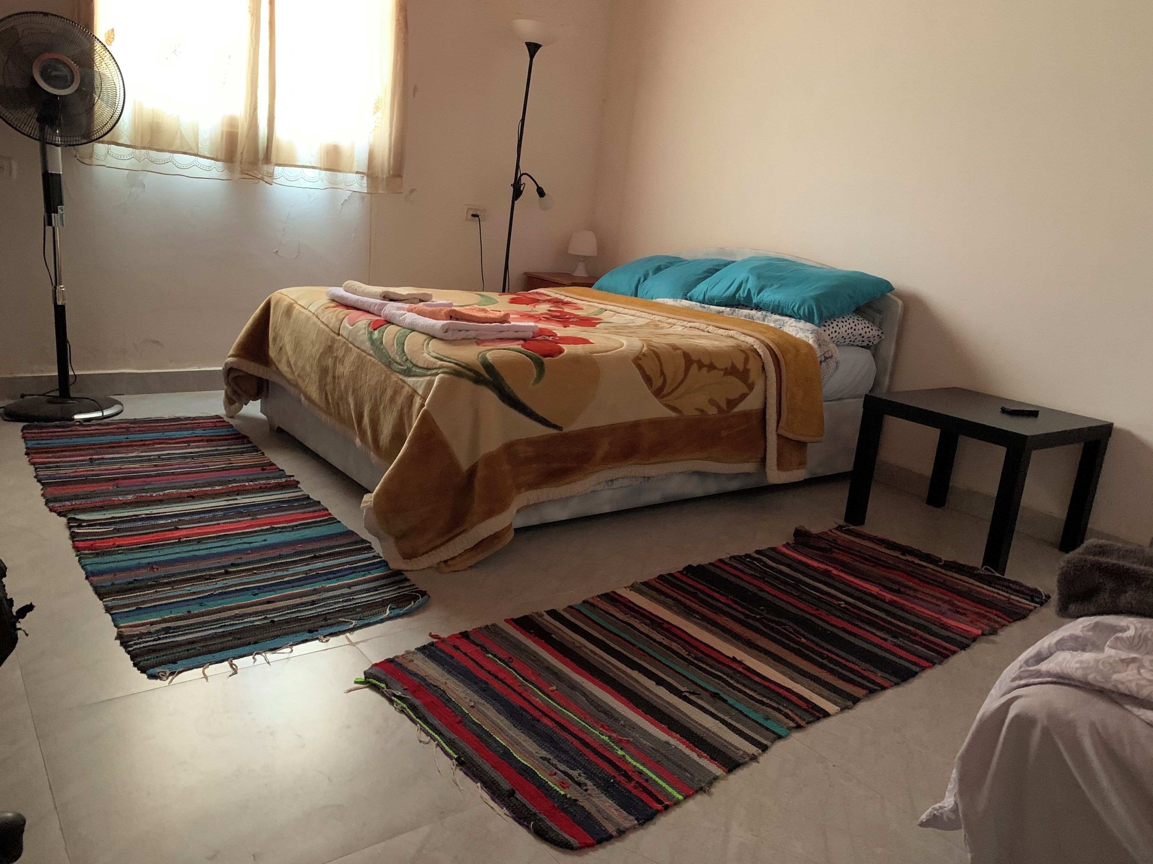 Un dormitorio con cama, dos ventiladores, una mesita de noche y dos alfombras de colores.
