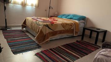 Quarto com cama, duas ventoinhas, mesa de cabeceira e dois tapetes coloridos.