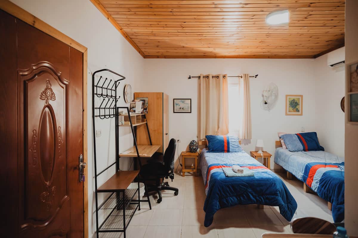 A room with a wooden ceiling, a desk with a chair, two beds with blue bedding, a wardrobe, and a door.