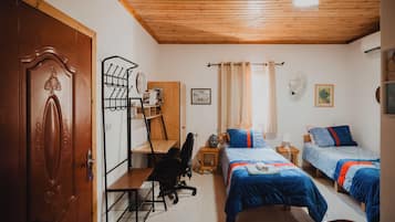 A room with a wooden ceiling, a desk with a chair, two beds with blue bedding, a wardrobe, and a door.