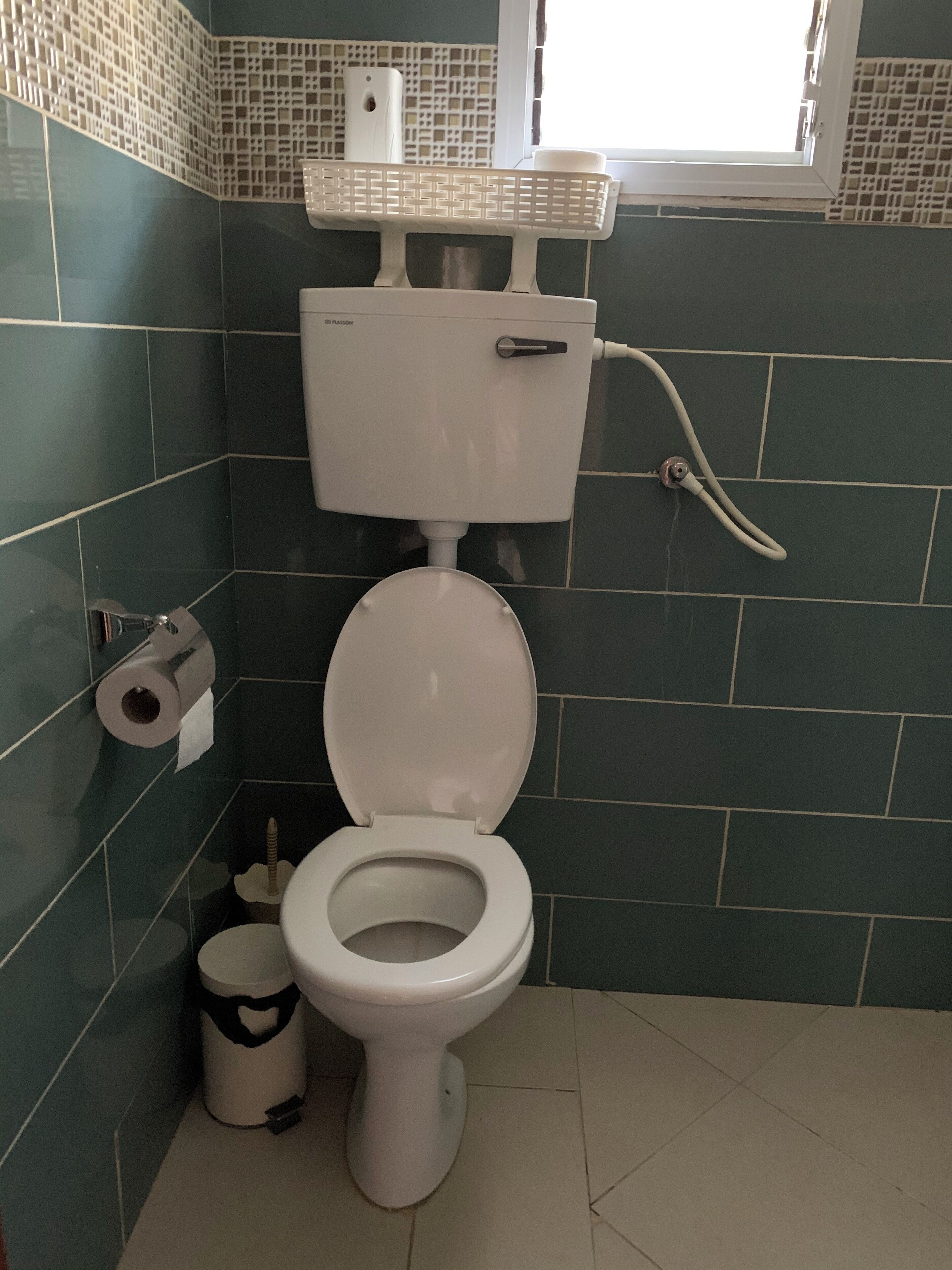 Een badkamer met een toilet, toiletrolhouder, prullenbak en een raam.