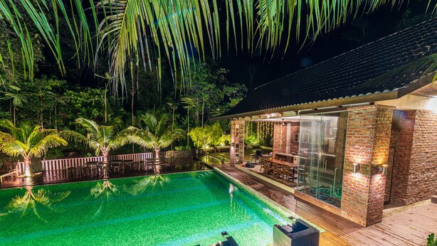 A nighttime view of a swimming pool with a wooden deck and a glass-enclosed patio area. - Bentong Eco Wellness Resort (Raub)