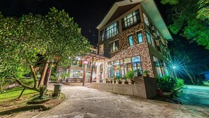 A multi-story building with a stone facade, large windows, and a well-lit entrance area with potted plants and a pathway leading to the door. - Bentong Eco Wellness Resort (Raub)