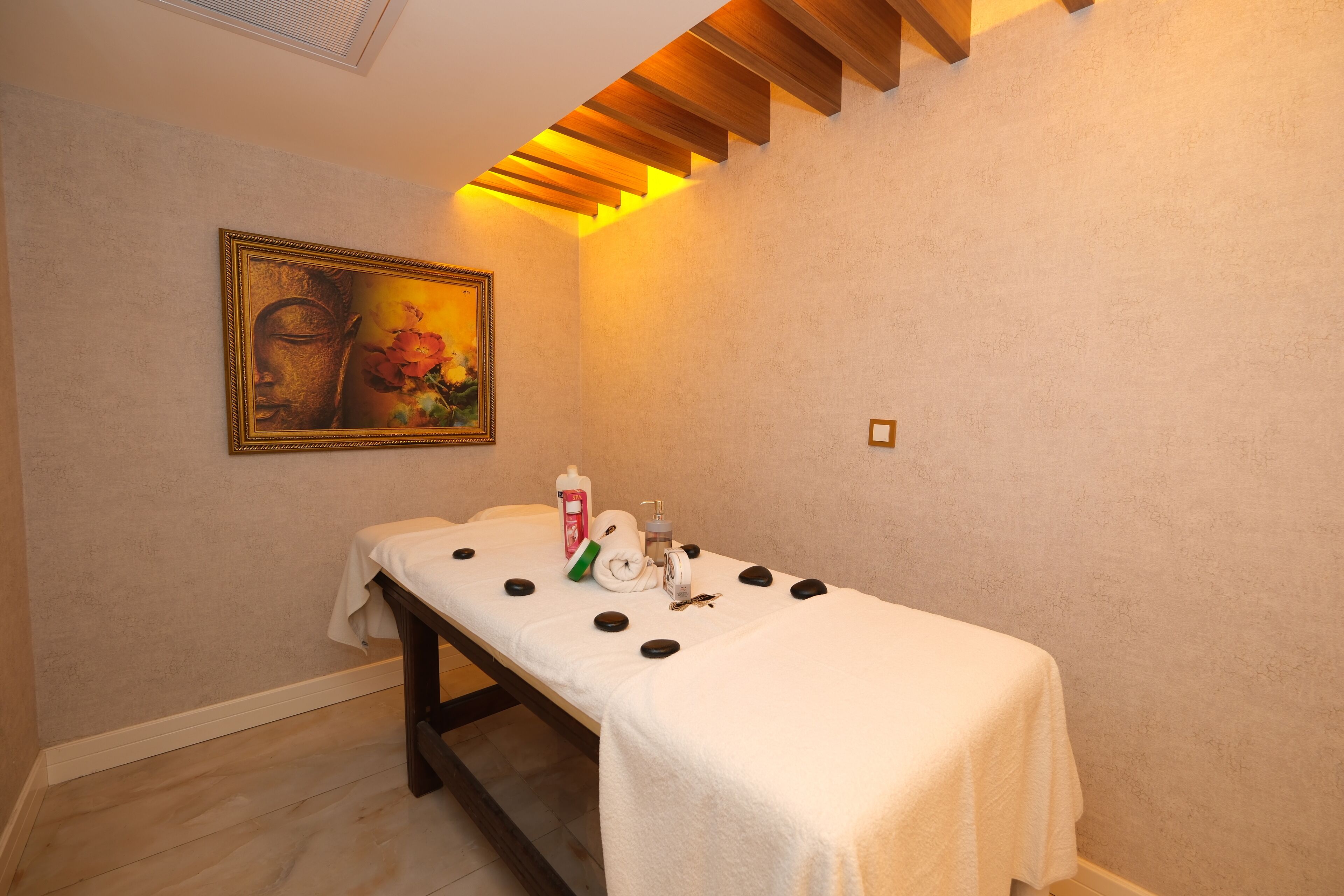 Ein Massagetisch mit weißem Handtuch, schwarzen Steinen und einer Ölflasche in einem Raum mit Holzboden und einem gerahmten Gemälde an der Wand.