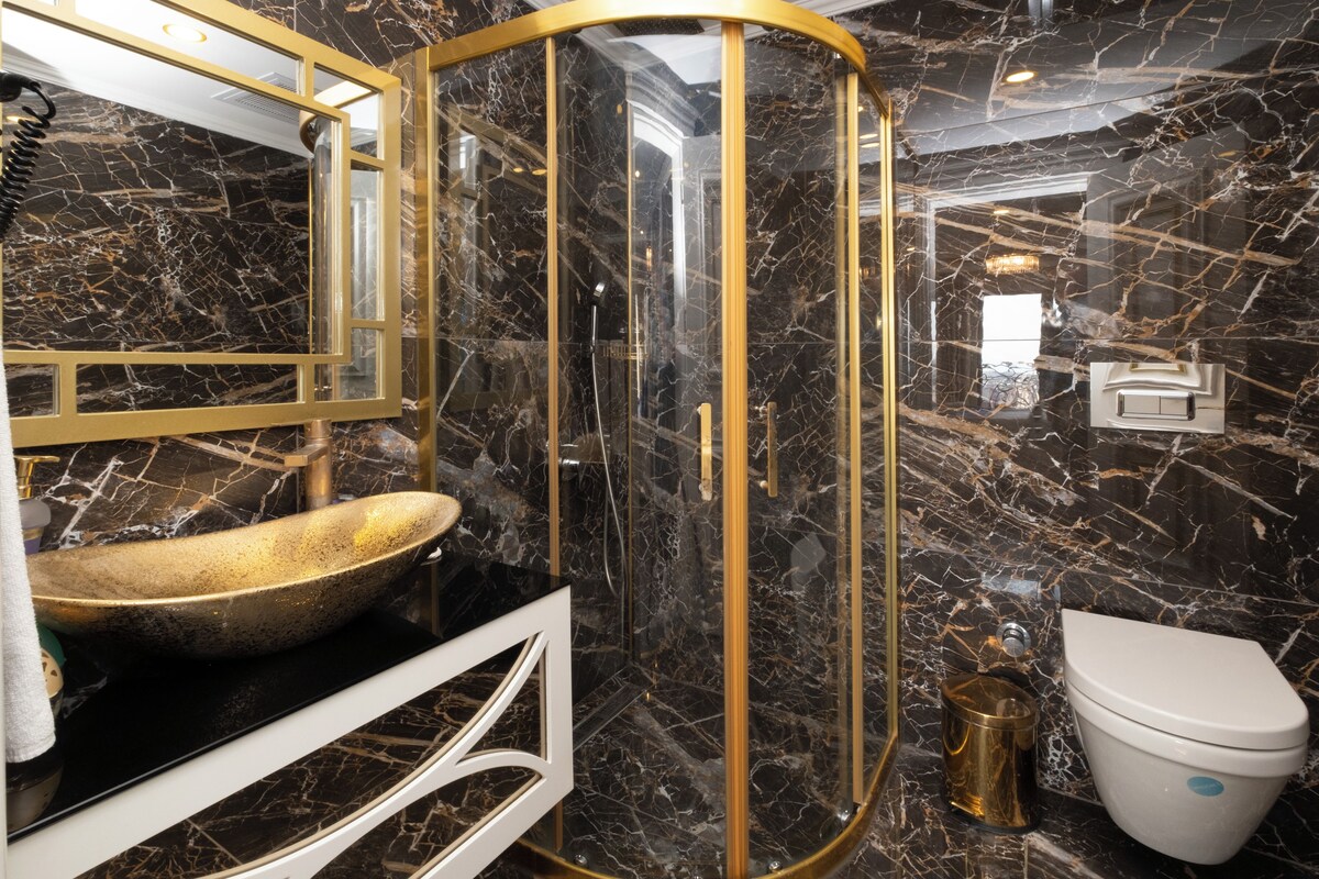 A bathroom with a gold sink, marble walls, and a gold-framed shower enclosure.