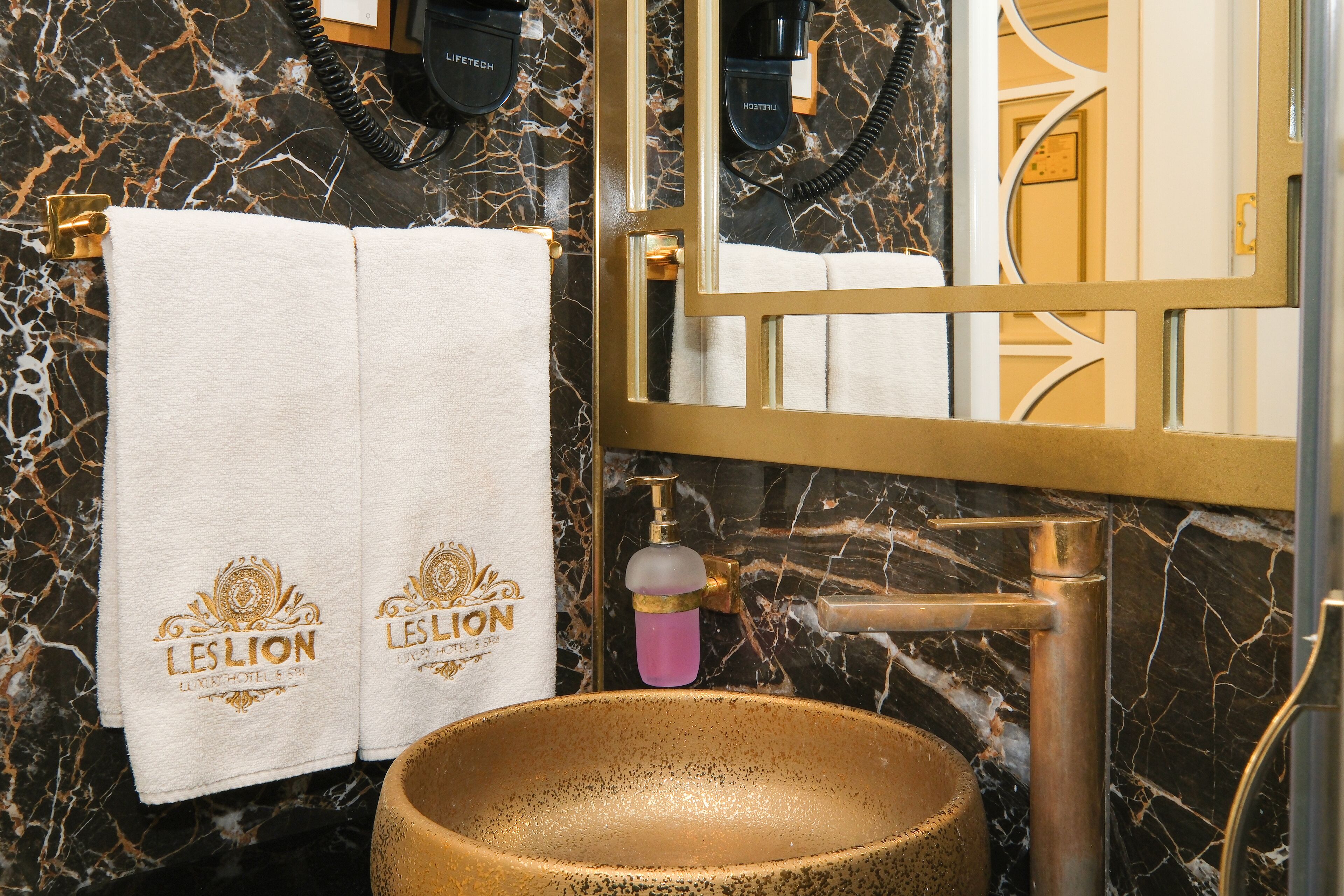 A bathroom with a marble wall, a gold sink, and two towels with 'LES LION' branding.
