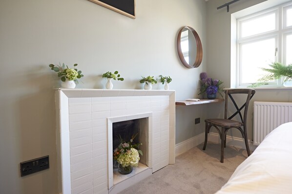 Una habitación con chimenea de baldosas blancas, una silla de madera, una mesita con un espejo y una ventana que da a zonas verdes.