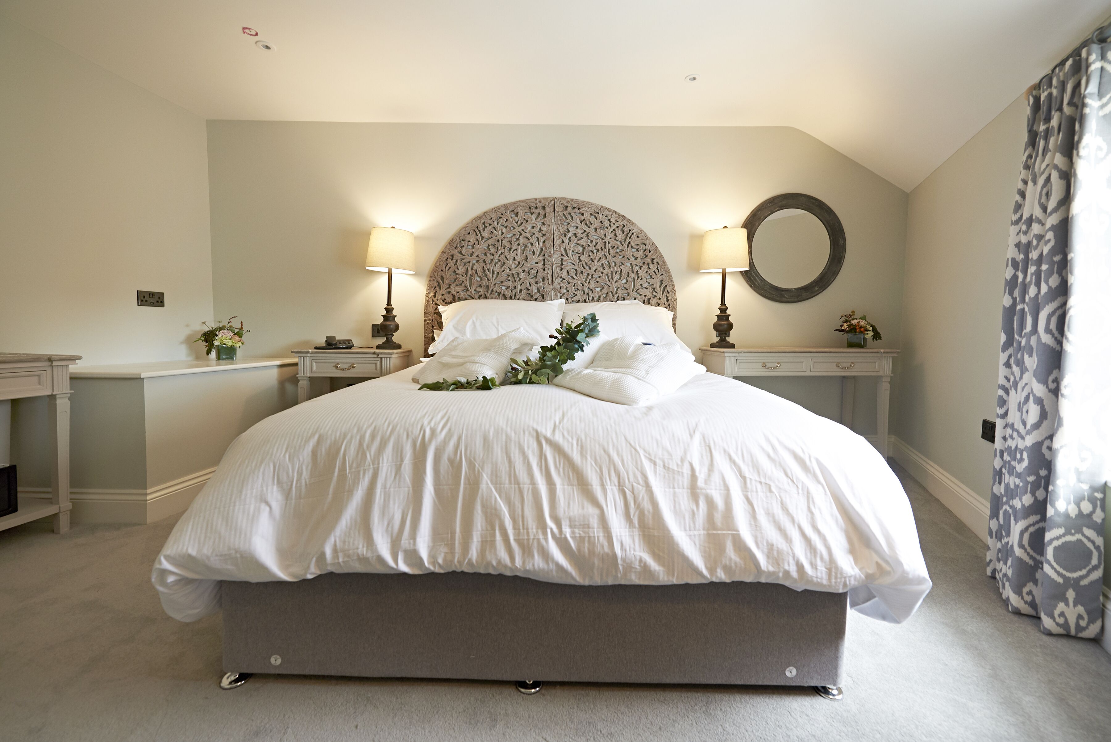 A bedroom with a large bed, two bedside tables, a round mirror, and a nightstand with a lamp.