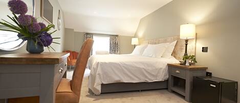 A modern hotel room with a bed, bedside tables, a desk with a chair, a small refrigerator, and a window with curtains.