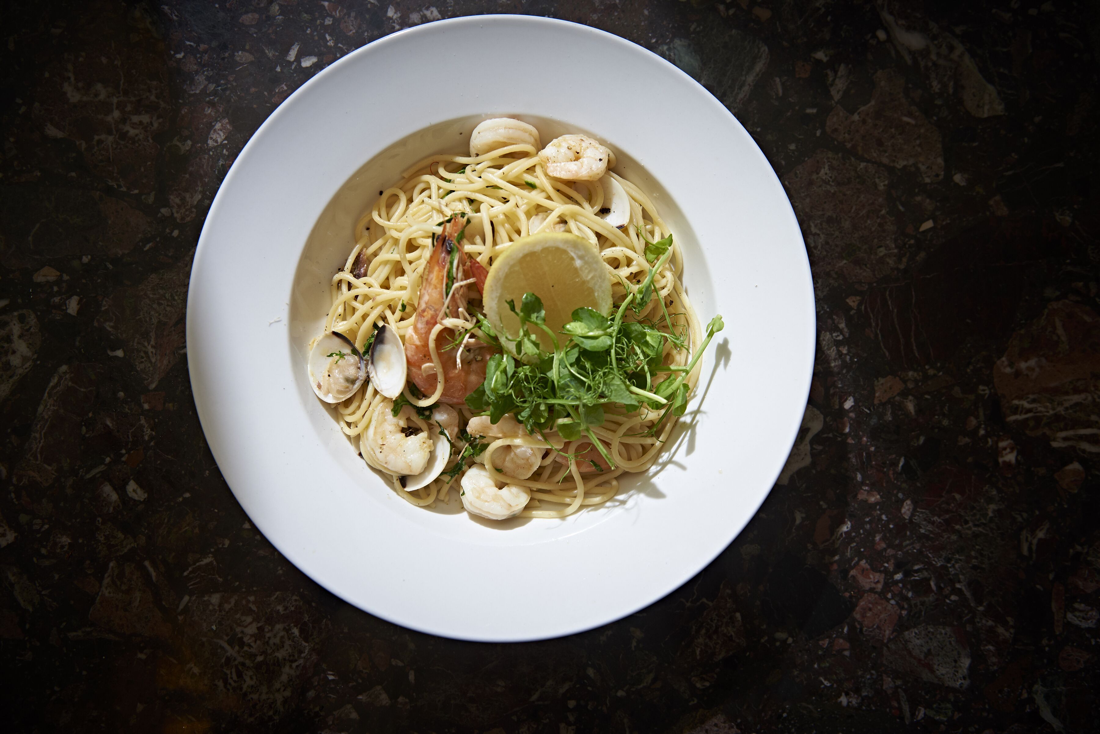 A plate of pasta with shrimp and clams, garnished with lemon and greens, on a dark stone surface.