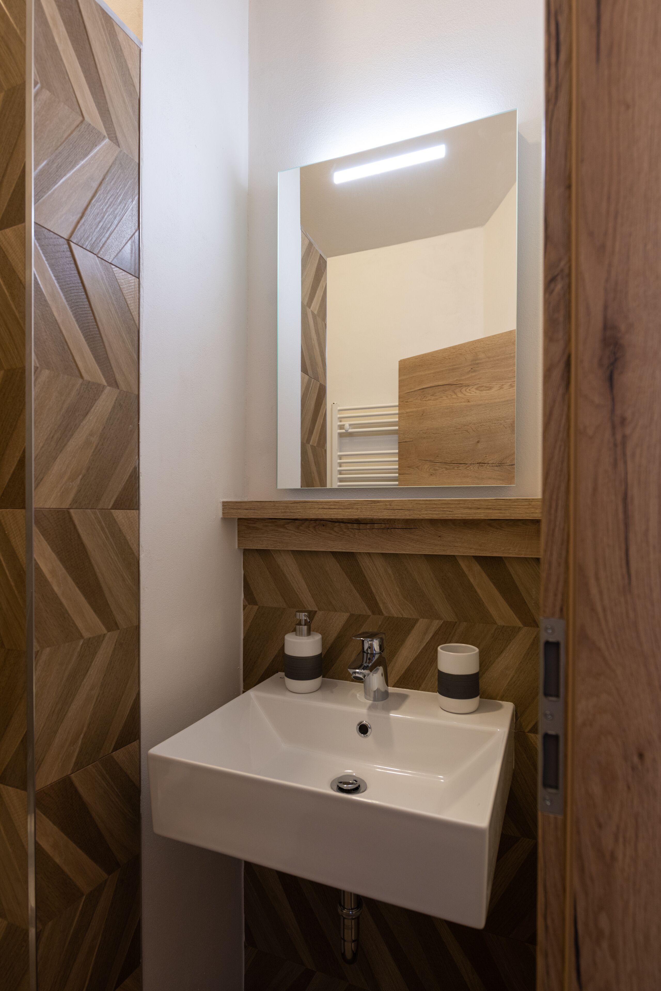 A bathroom with a white sink, two soap dispensers, a mirror, and a wooden wall with a herringbone pattern.