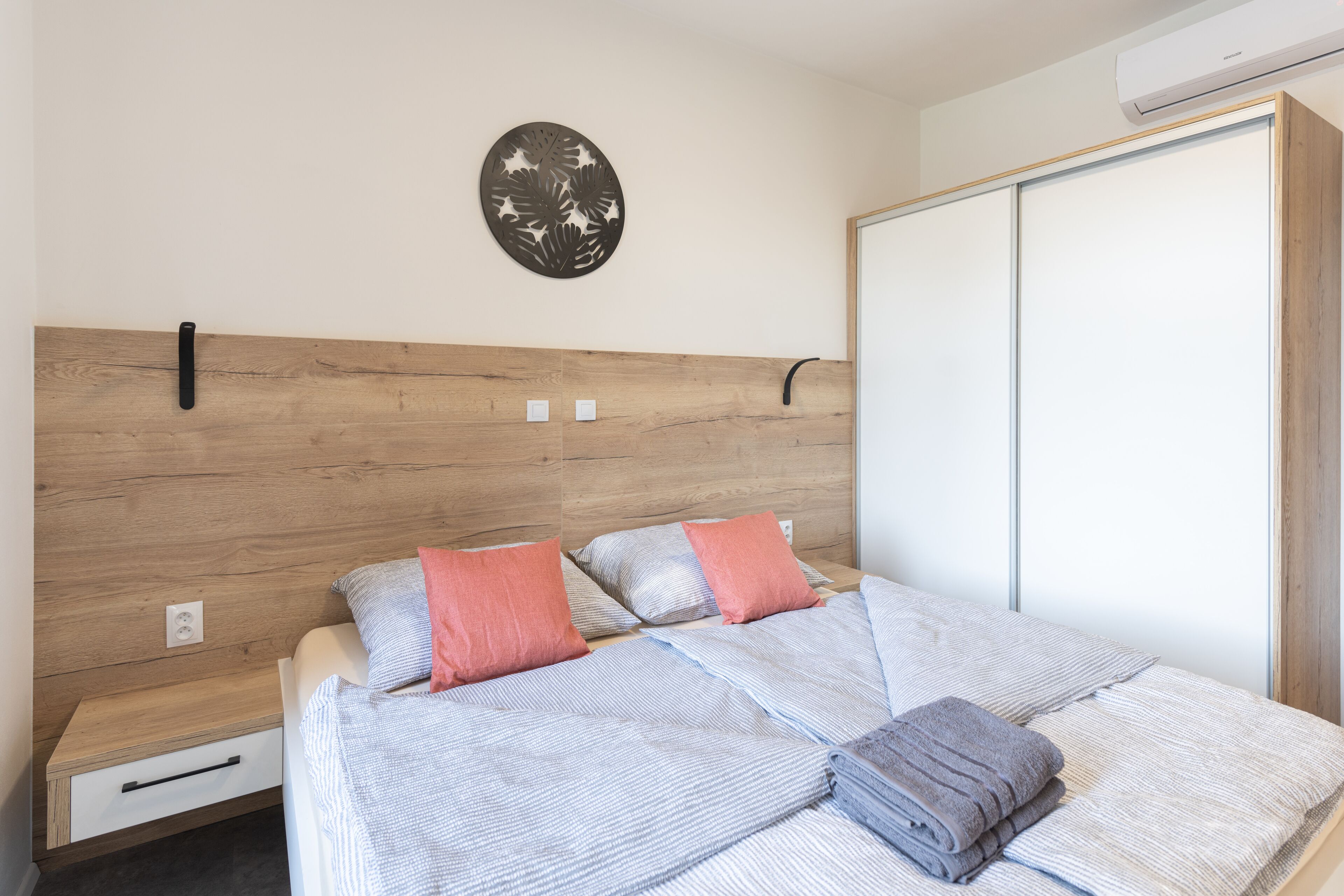 A bedroom with a wooden headboard, a bed with striped bedding, two pillows, a folded towel, a nightstand with a drawer, and a wall-mounted decorative piece.