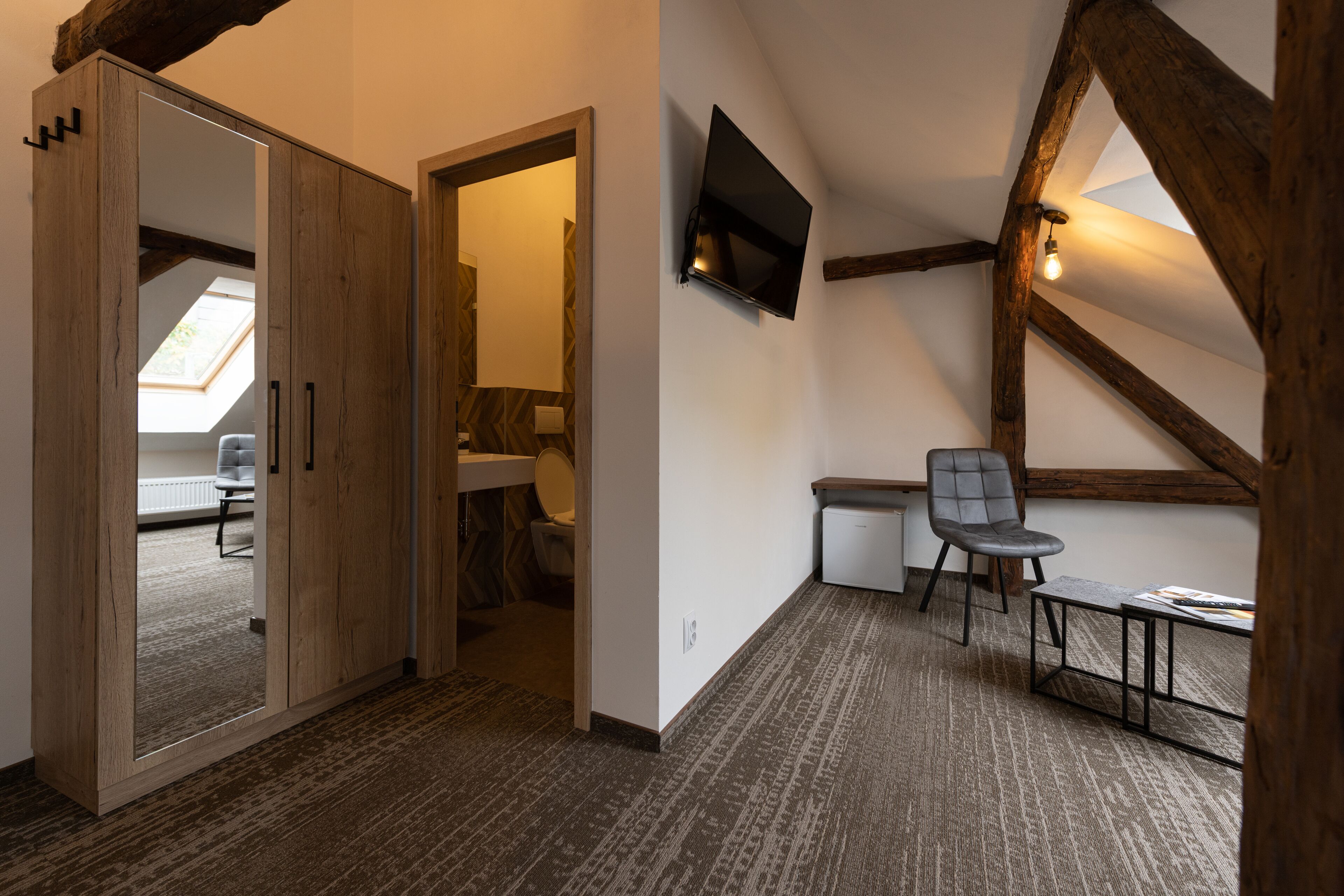Una stanza con soffitto spiovente, travi in legno, una TV a schermo piatto montata al muro, una poltrona grigia, un tavolino e pavimento rivestito di moquette.