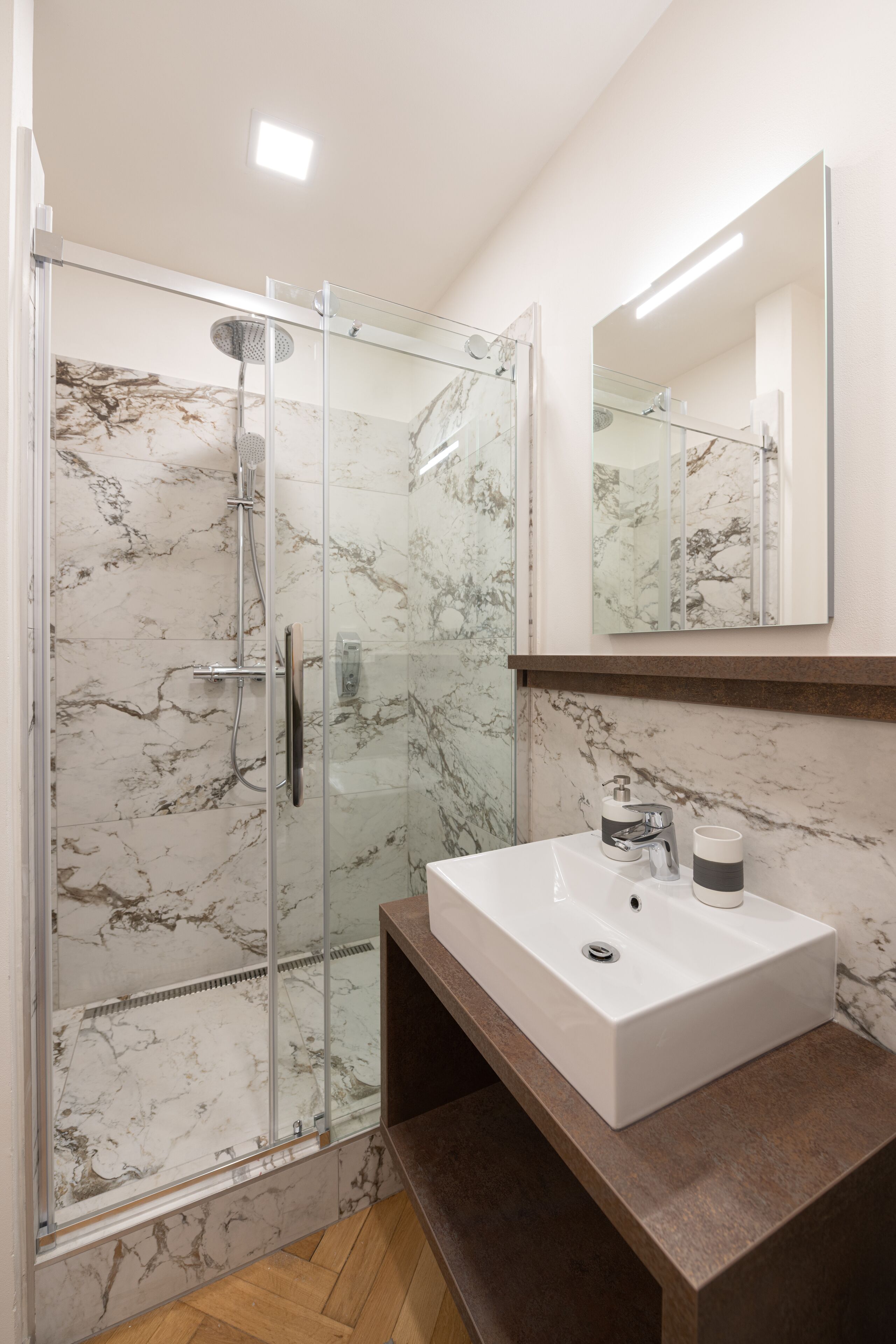 Un baño moderno con ducha de vidrio, paredes de mármol, un lavabo blanco y un espejo grande.