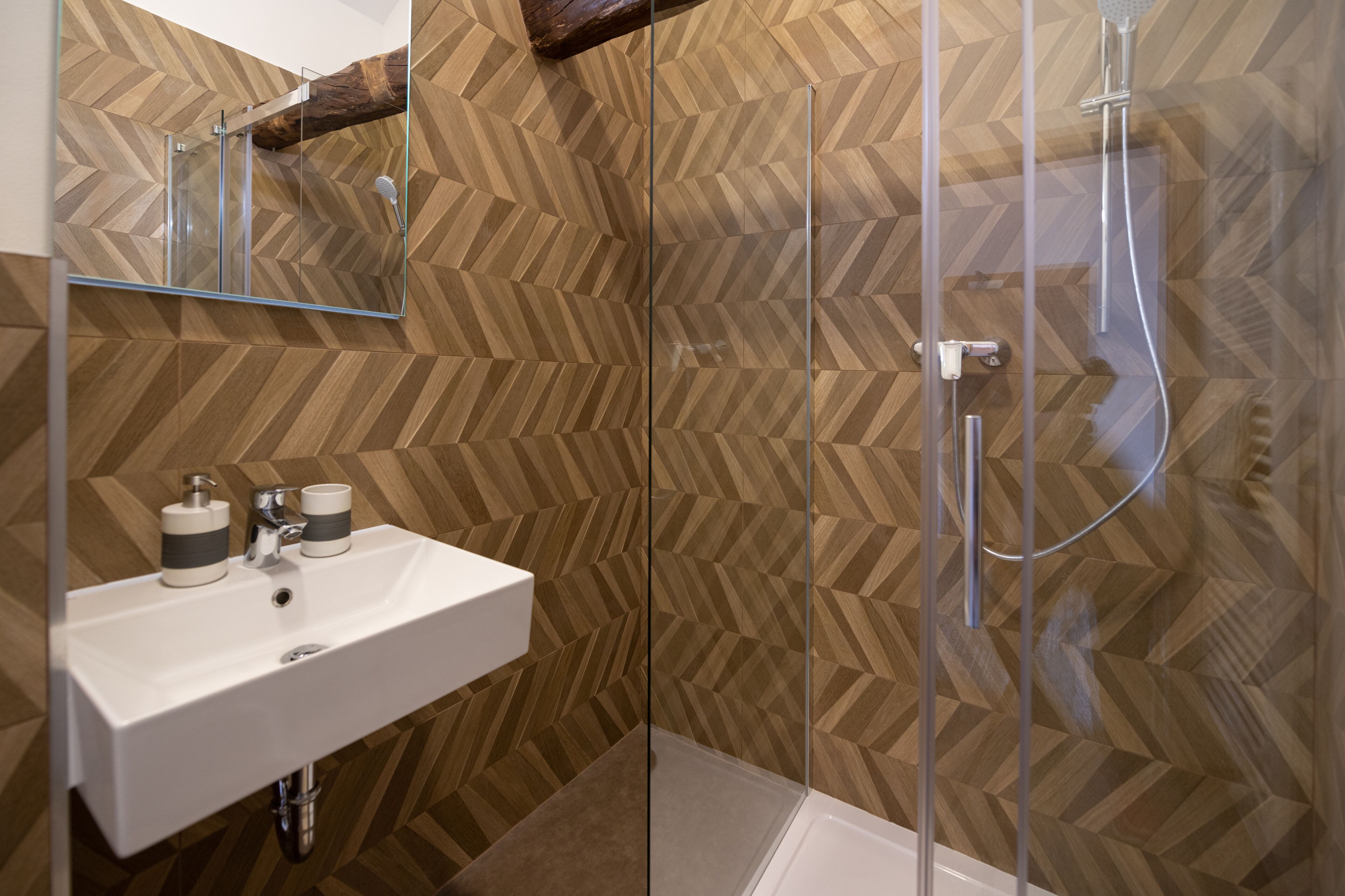 A modern bathroom with a wooden herringbone pattern wall, a glass shower enclosure, and a white sink with a mirror above it.