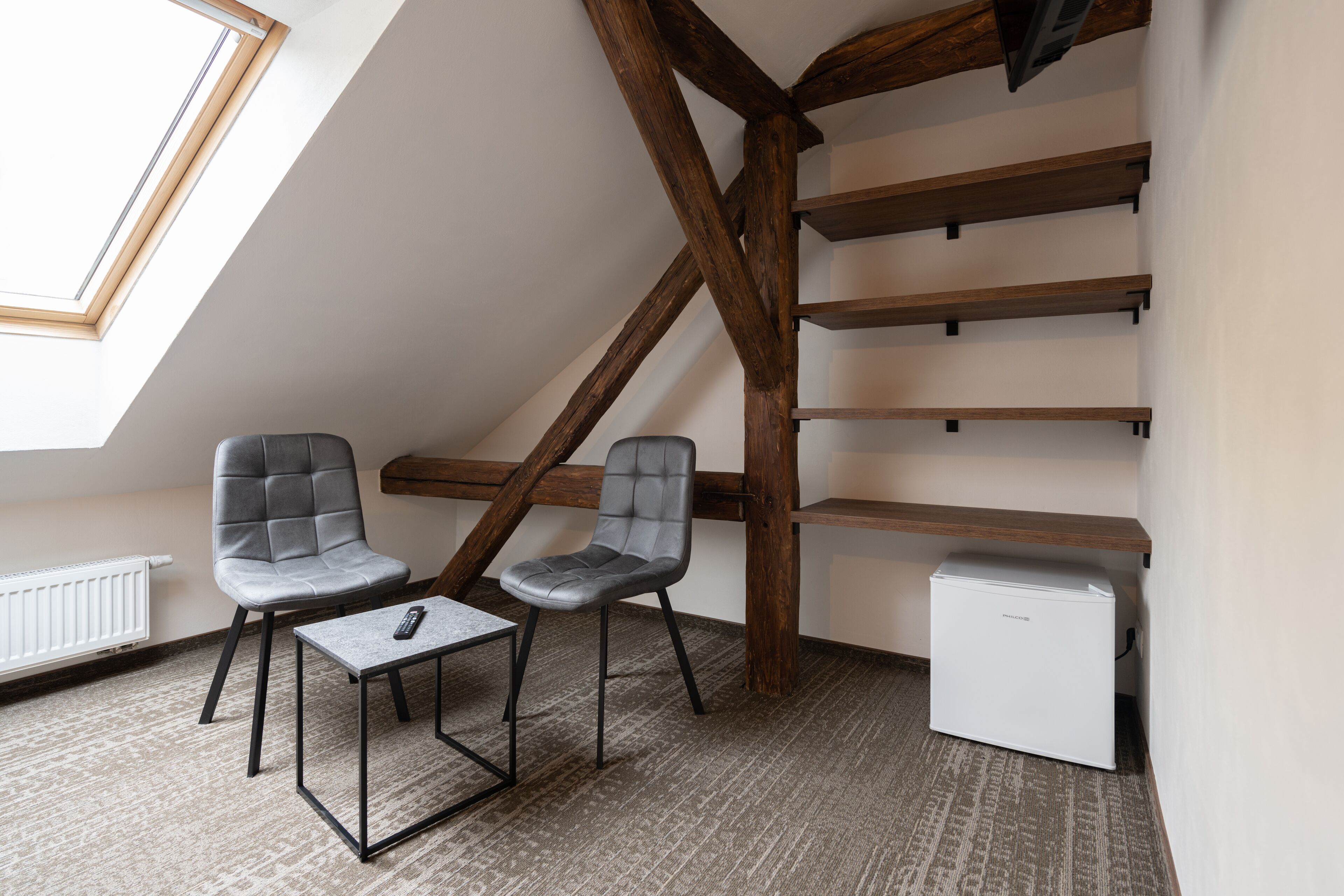A small, modern room with a sloped ceiling, two grey upholstered chairs, a small table with a remote control, a white mini-fridge, and wooden beams.