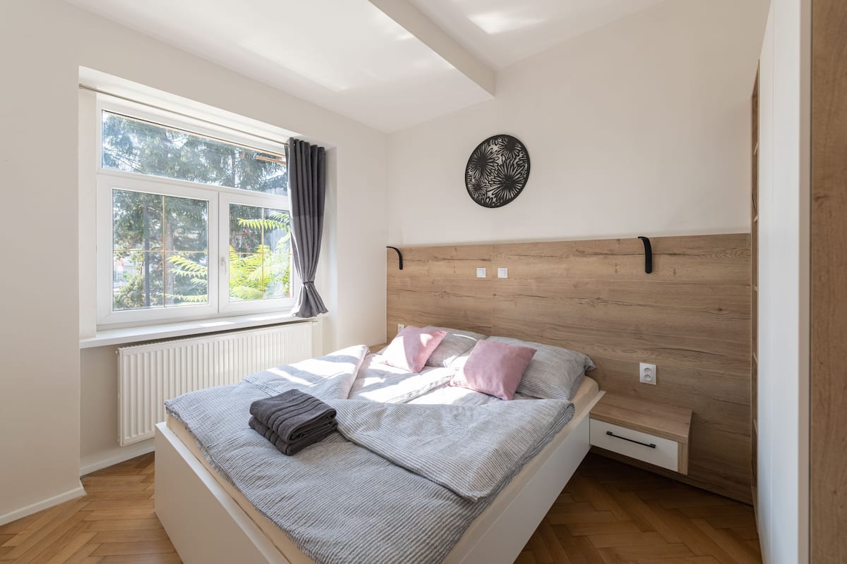 A bedroom with a wooden headboard, a large window with curtains, a radiator, and a bed with pillows and a blanket.