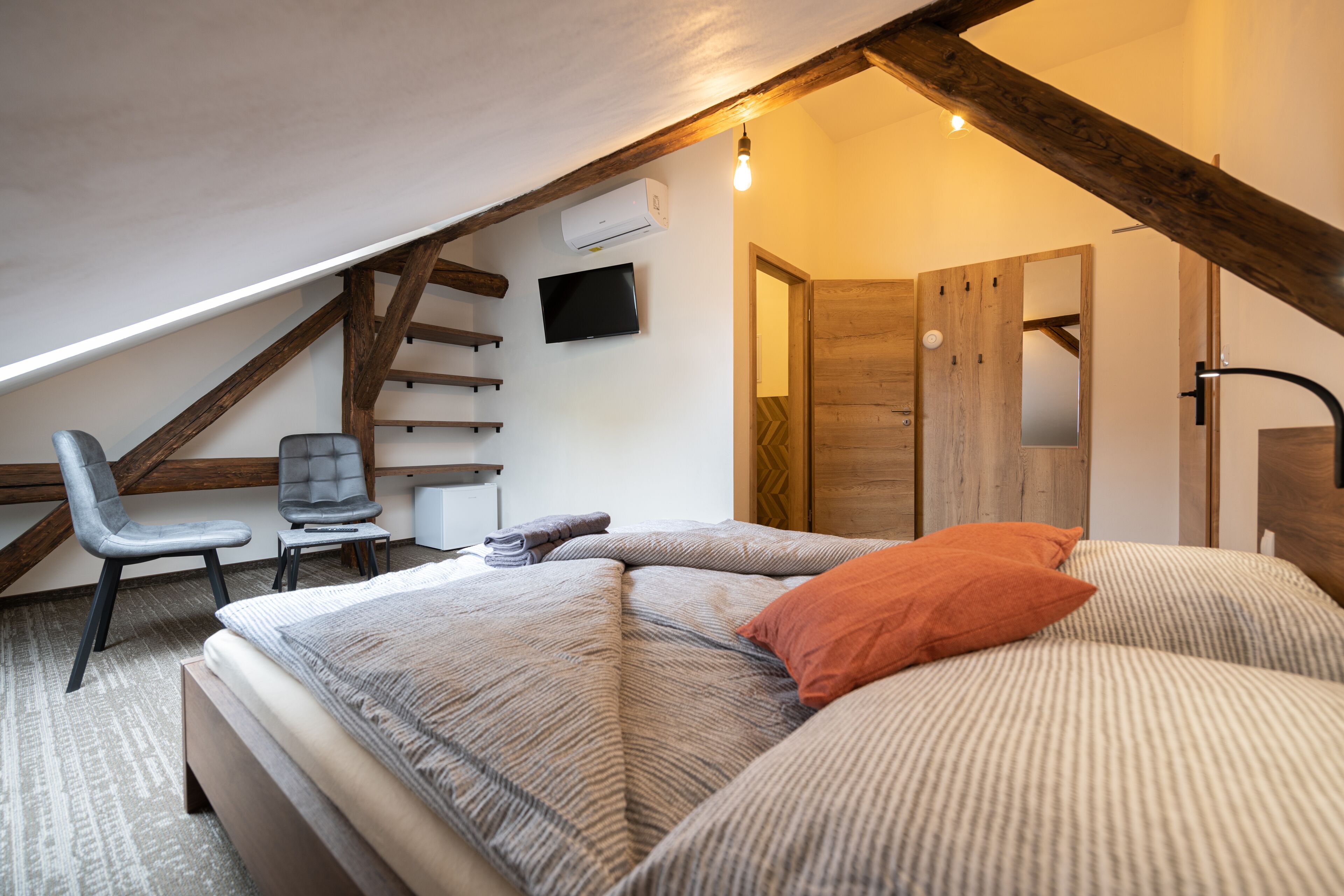 A bedroom with a sloped ceiling, a bed with striped bedding, a chair, a flat-screen TV, and a wooden wardrobe.