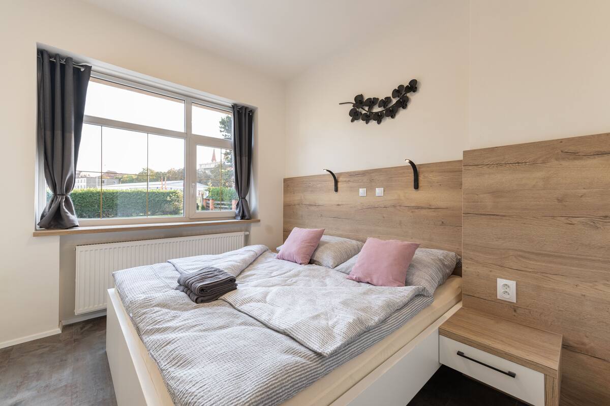 A bedroom with a bed, a window with curtains, a radiator, a wall-mounted light, and a small bedside table.