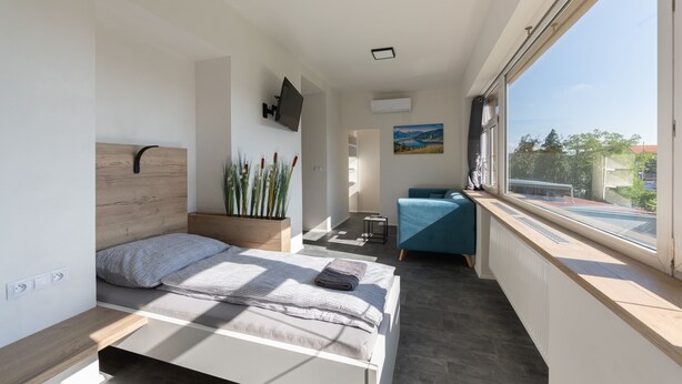A modern bedroom with a bed, bedside table, potted plants, a view of the outdoors, and a window. - Apartsee Wellness Plzeň (Plzen)