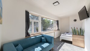 A modern living room with a blue sofa, a white bed, a large window, and a mounted TV. - Apartsee Wellness Plzeň (Plzen)