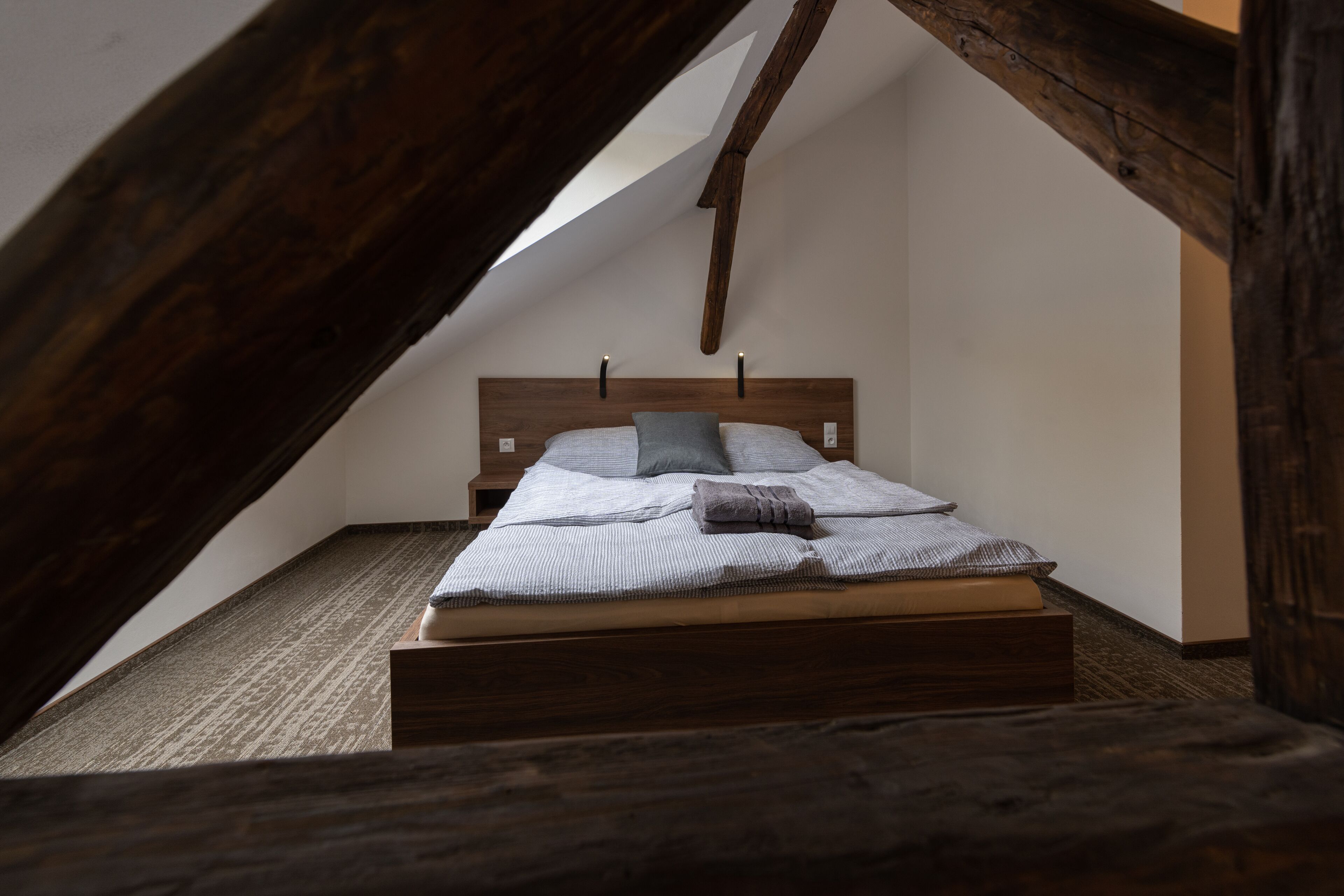 A bedroom with a sloped ceiling, wooden beams, a bed with a headboard, and a small bedside table.