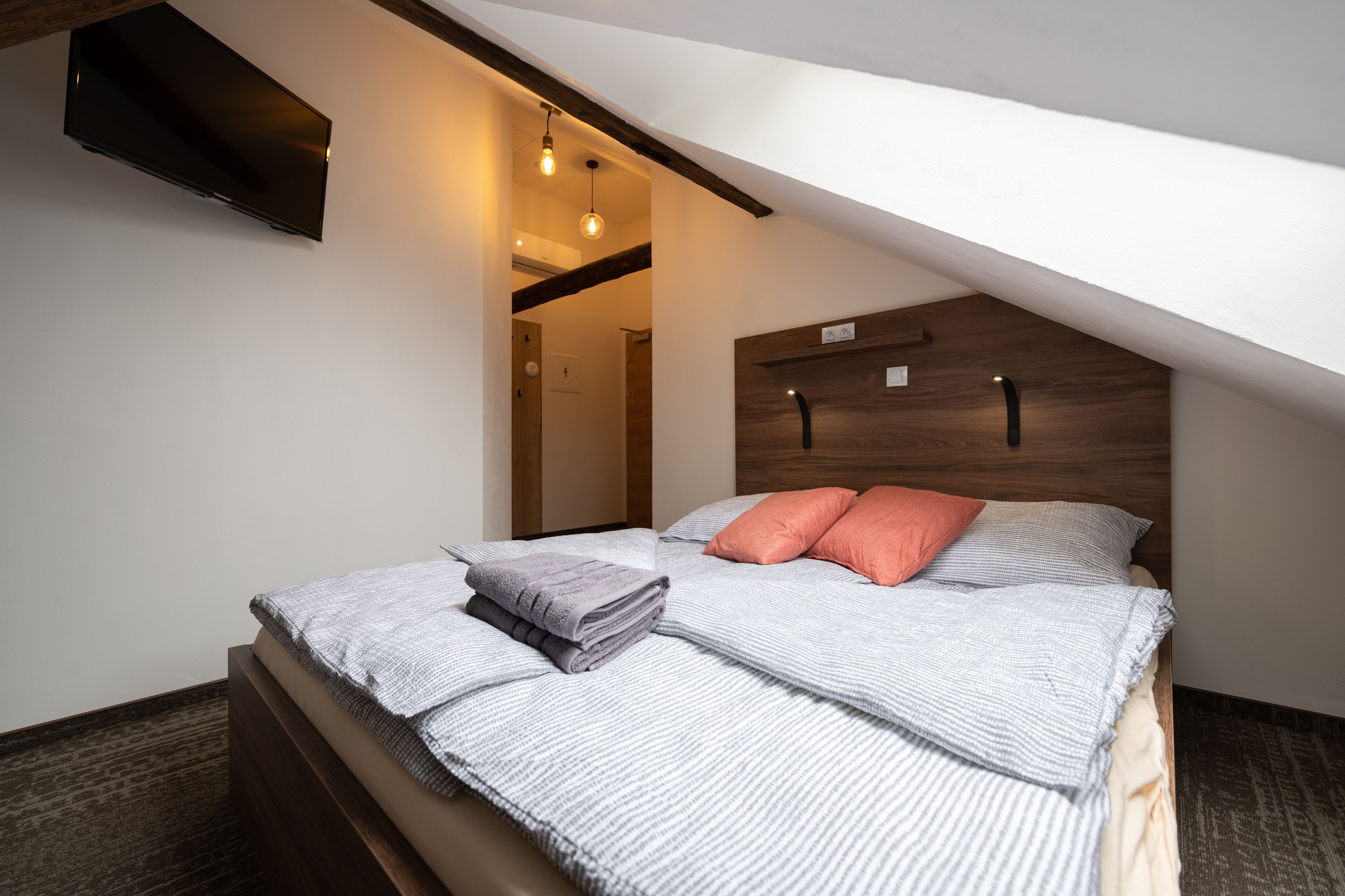Une chambre moderne avec un plafond en pente, un lit doté d’une tête de lit en bois et un petit téléviseur fixé au mur.