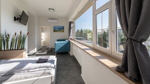 A modern hotel room with a large window, a bed, a desk, and a view of the city. - Apartsee Wellness Plzeň (Plzen)